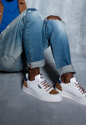 Persoon die gescheurde blauwe spijkerbroek en witte sneakers met bruine suède accenten draagt, zittend op een grijze vloer.