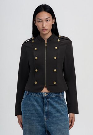 Donna con capelli lunghi e neri che indossa una giacca corta nera con bottoni dorati e jeans blu a vita alta, rivolta frontalmente su uno sfondo semplice.