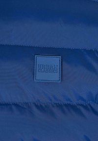 Marineblå pufferjakke i materiale med en glat overflade. Dækker en firkantet logo-patch mærket "URBAN CLASSICS" i midten.