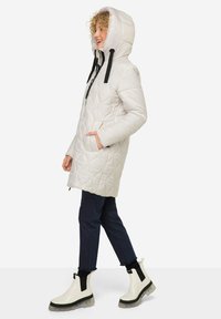 Parka blanche matelassée avec une capuche, cordons de serrage noirs, deux poches latérales, associée à un jean foncé et des bottines blanches à semelles épaisses.