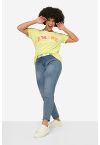 Camiseta amarilla de manga corta con la palabra "AMOUR" en letras rosas y naranjas, combinada con unos jeans ajustados azules y zapatillas blancas.