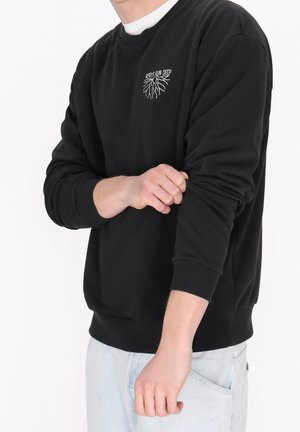 Personne remontant la manche d’un sweat-shirt noir avec l’inscription « Roots Run Deep » et un motif de racine sur la poitrine, portant un jean bleu clair.