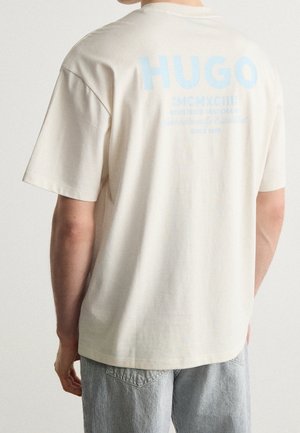 Osoba nosząca kremowy T-shirt z dużym jasnoniebieskim napisem "HUGO" na plecach i szare spodnie w paski, stojąca tyłem.