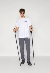 Νεαρός άνδρας με γυαλιά ηλίου, λευκό μπλουζάκι Terrex, γκρι παντελόνι, κρατώντας μαύρα μανίκια πεζοπορίας, διασφαλίζοντας μπροστά από έναν απλό φωτεινό φόντο.