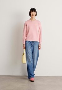 Vrouw in een roze sweatshirt, blauwe jeans en roze schoenen, met een lichtgele handtas, staande tegen een effen witte muur.