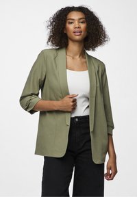 Olivgrön blazer med avslappnad passform, uppvikt ärmar och mjuk textur, bärs över en vit topp och matchas med svarta byxor.