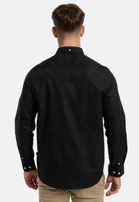 Muž otočený chrbtom, oblečený v čiernej košeli s dlhým rukávom a béžových nohaviciach, na jednoliatom svetlom pozadí.