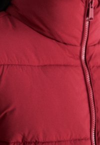 Sekcja czerwonej kurtki puchowej z widocznym poziomym przeszyciem i częściowo widocznym pasującym czerwonym zamkiem błyskawicznym.