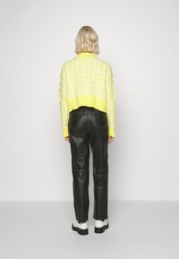 Person med kort blont hår som bär en gul stickad tröja, svarta läderbyxor och vita kängor med tjock sula, står vänd mot en enkel vit vägg.