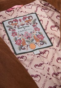 Hnědá kordurová tkanina s růžovou podšívkou, která obsahuje květinové a zvířecí vzory. Štítek zobrazuje "Barbour" a "FARM RIO" v dekorativním písmu.