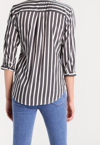 Camicia a righe grigie e bianche con maniche a tre quarti, orlo arrotondato e dettagli con bottoni ai polsini. Indossata con jeans blu.
