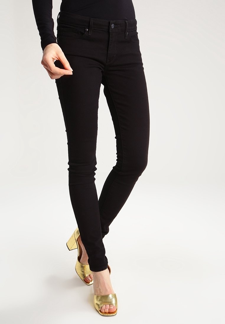 Personne portant un jean noir slim et des chaussures à talons hauts ouvertes dorées, debout avec les jambes croisées.