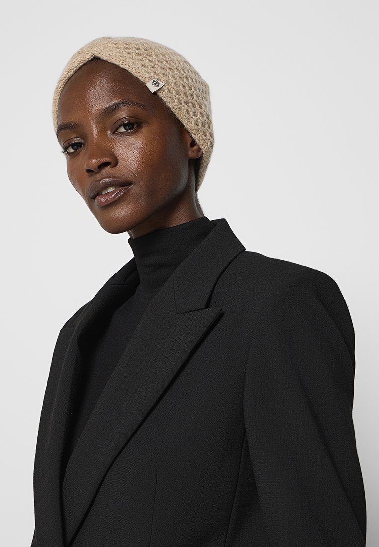 Beige Strickmütze mit strukturiertem Muster; getragen mit einem schwarzen Rollkragenpullover und Blazer. Verfügt über ein kleines Markenschild an der Seite.