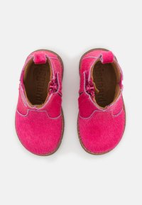 Chaussures pour enfants de couleur fuchsia, fabriquées à partir d'un matériau doux et texturé avec une semelle marron. Présentent une couture décorative et un petit onglet sur le talon.