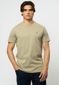 Lys beige kortermet T-skjorte, rund hals, myk tekstur, med en liten grønn logo på brystet. Brukt med mørkeblå jeans.