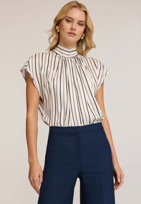 Blusa crema con strisce verticali blu navy, collo alto e maniche corte. Abbinata a pantaloni navy su misura, con texture liscia e drappeggio delicato.