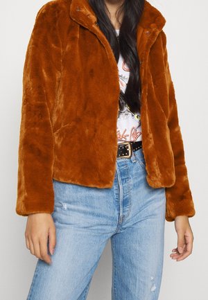 Frau trägt eine rostfarbene Kunstpelzjacke, weißes grafisches T-Shirt, schwarzen Nieten-Gürtel und hellblaue hoch taillierte Jeans.
