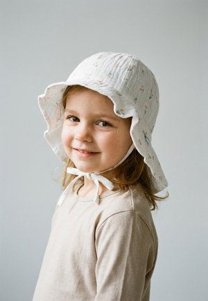 Lächendes junges Mädchen mit hellbraunen Haaren, das ein beiges Langarmshirt und einen weißen Sonnenhut mit Blumenmuster trägt, der unter dem Kinn gebunden ist.