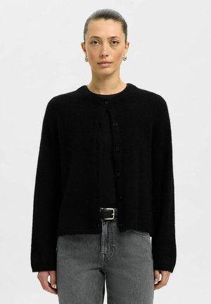Schwarze Strickjacke aus weichem, flauschigem Material; runder Halsauschnitt, Knopfverschluss, lange Ärmel. Kombiniert mit einem schwarzen Oberteil und grauen, hochgeschnittenen Jeans.