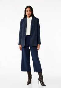 Marineblauwe pinstripe blazer over een wit polo, gecombineerd met marineblauwe wijde broek en bruine stilletto laarzen, tegen een witte achtergrond.