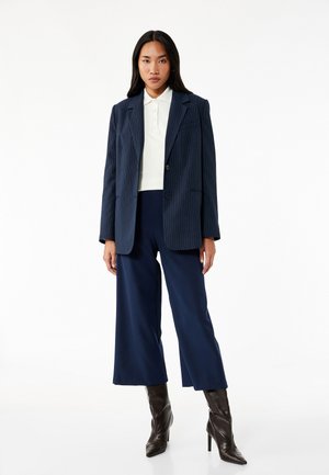 Marineblauer Nadelstreifen-Blazer über einem weißen Polo, kombiniert mit marineblauen weiten Hosen und braunen Stiletto-Stiefeln, vor einem weißen Hintergrund.