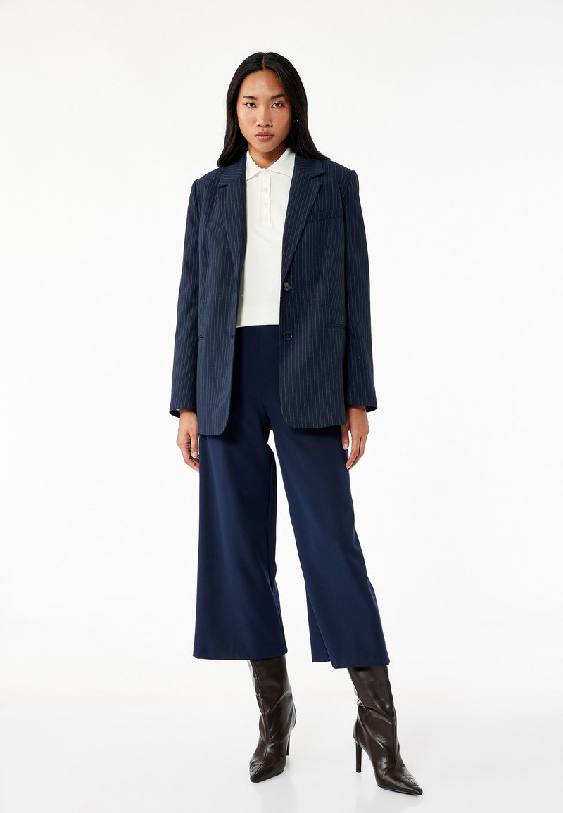 Marineblauwe pinstripe blazer over een wit polo, gecombineerd met marineblauwe wijde broek en bruine stilletto laarzen, tegen een witte achtergrond.