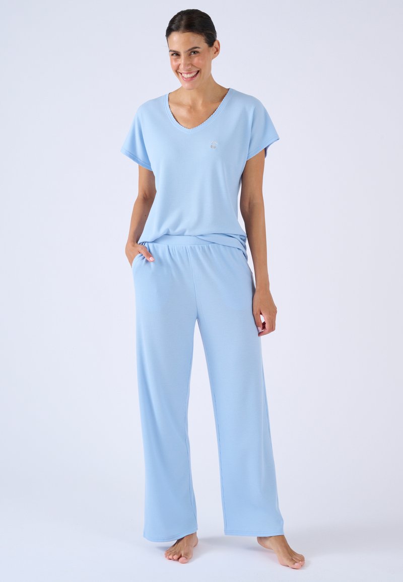 Femme souriante, debout pieds nus, vêtue d'un haut à manches courtes bleu clair et d'un pantalon ample assorti, les mains dans les poches sur un fond uni.