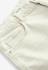 Pantalon en coton beige avec une texture lisse, présentant une poche arrière et un détail de bouton en métal près de la taille. Design épuré et minimaliste.