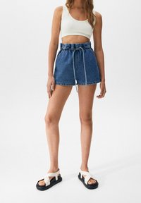 Pantalones cortos de mezclilla con cintura elástica y cordón en azul. Combinados con una camiseta corta de canalé blanca y sandalias negras con tiras blancas.