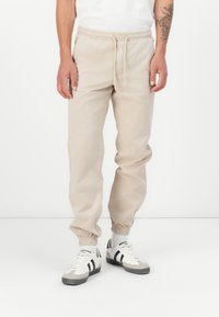 Beige cotton joggers with elastic waistband and cuffs. Features side pockets and a relaxed fit. Paired with white sneakers and gray accents.