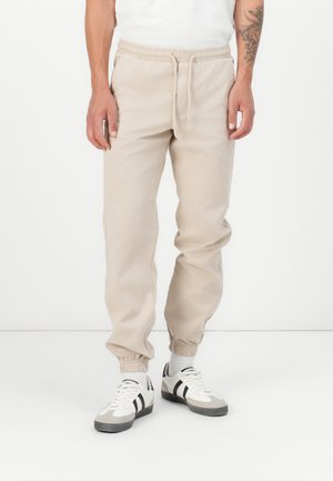 Joggers de algodón beige con cintura y puños elásticos. Cuenta con bolsillos laterales y un corte relajado. Combinados con zapatillas blancas y acentos grises.