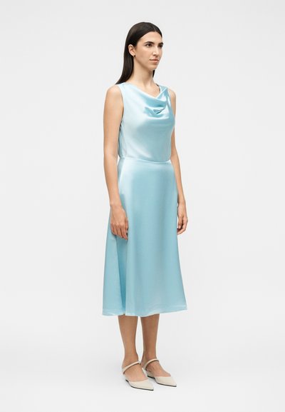 Γυναίκα που φοράει φωτεινό γαλάζιο σατέν μίντι φόρεμα χωρίς μανίκια με βολάν στο ντεκολτέ και μπεζ μυτερά παπούτσια σε λευκό φόντο.