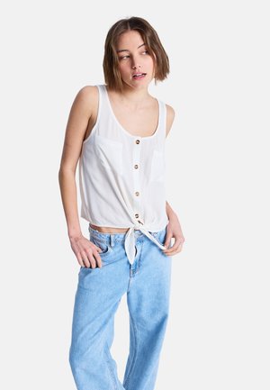 Witte, lichtgewicht mouwloze top met knoopsluiting aan de voorkant en twee borstzakken, gestrikt in de taille. Gecombineerd met lichtblauwe wijde jeans.