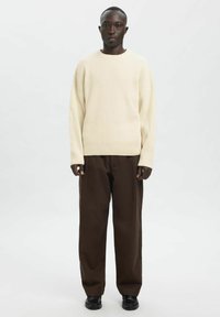 Beežist soonikkoes kudum, mida on kombineeritud lahtiste tumepruunide pükstega ja mustade kingadega, valge ühevärvilise taustaga.