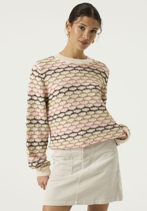Mujer lleva un jersey claro con líneas geométricas y una falda beige clara, de pie con una mano en el bolsillo, sobre un fondo liso.