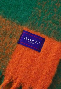 Gros plan sur un tissu en laine doux vert et orange avec une étiquette violette GANT Sportswear cousue sur la section orange.