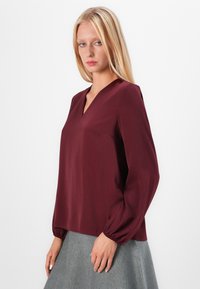 Burgunderfarget langermet bluse med V-hals, myk tekstur og rynkede erm cuffs, kombinert med en lys grå skjørt. Minimalistisk design og snitt.