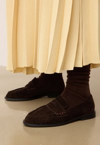 Mocassins en daim marron avec une texture lisse et des détails de couture subtils. Associés à des chaussettes côtelées marron foncé et une jupe plissée jaune clair.