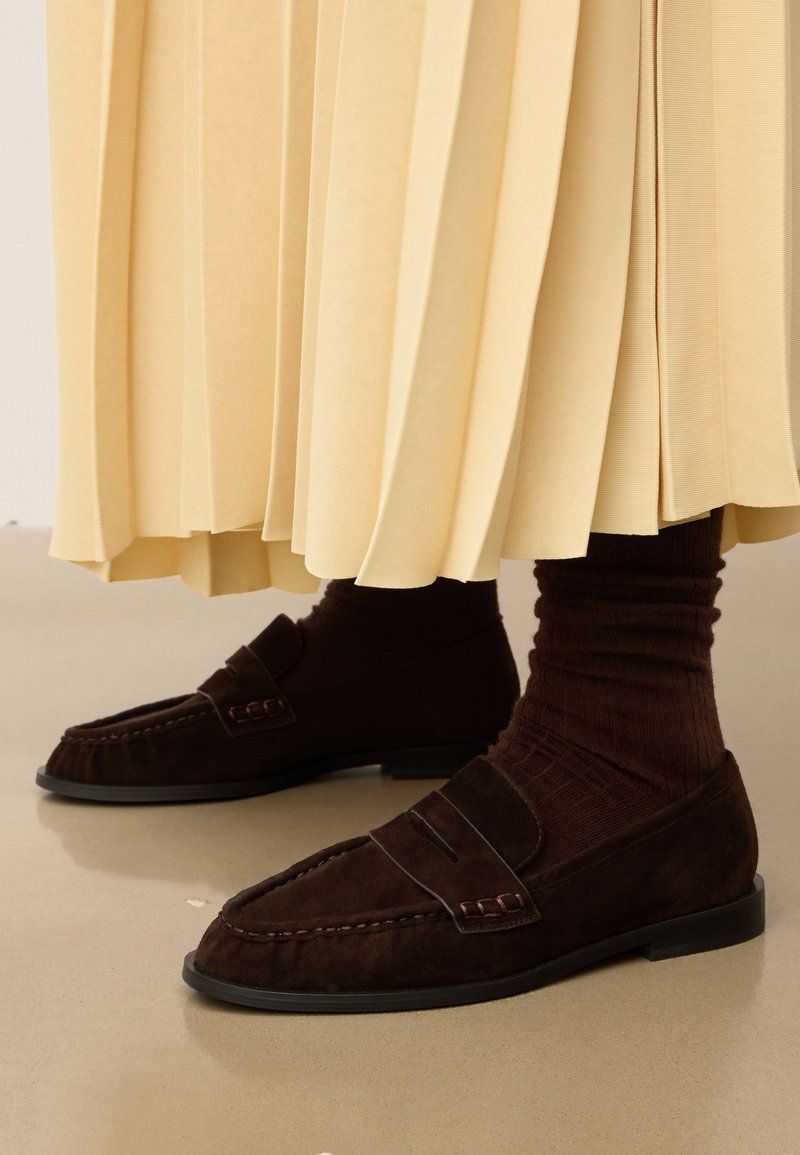 Mocassins en daim marron avec une texture lisse et des détails de couture subtils. Associés à des chaussettes côtelées marron foncé et une jupe plissée jaune clair.