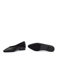 Zapatos planos negros de cuero para mujer con punta puntiaguda y tacón bajo en bloque, un zapato de pie y el otro con la suela hacia arriba.