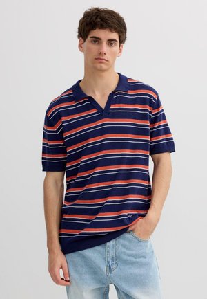 Joven con cabello oscuro que lleva una camiseta polo azul marino con rayas horizontales rojas y blancas y vaqueros azul claro, de pie con una mano en el bolsillo.