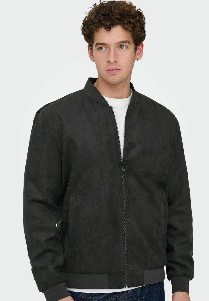 Jeune homme aux cheveux bouclés portant une veste bomber noire zippée par-dessus une chemise blanche, regardant sur le côté avec une expression neutre.