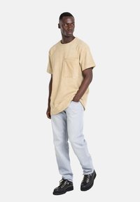 Beige Baumwoll-T-Shirt mit Brusttasche, kombiniert mit hellblauen Jeans und schwarzen Schnürstiefeln mit einer robusten Sohle.