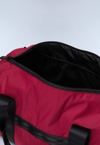 Sac en tissu rouge avec une fermeture éclair noire, ouverture large, doublure noire et deux poignées noires. L'intérieur dispose d'une poche en maille pour l'organisation.