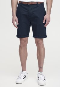 Navy kék pamutzshort, felgöngyölt szárakkal, barna bőrövet viselve. Fehér sportcipővel, fekete kiegészítőkkel párosítva. Tiszta dizájn.