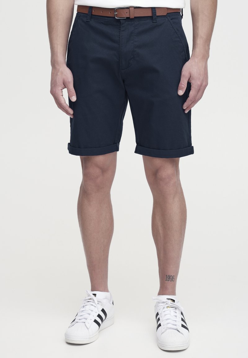Navy kék pamutzshort, felgöngyölt szárakkal, barna bőrövet viselve. Fehér sportcipővel, fekete kiegészítőkkel párosítva. Tiszta dizájn.