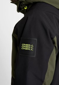 Personne portant une veste noire et olive avec un patch sur la manche montrant "O'NEILL" répété trois fois en texte jaune.