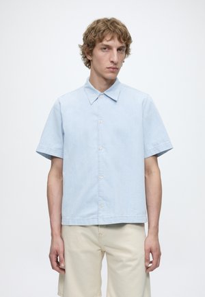 Jeune homme aux cheveux bouclés portant une chemise bleu clair à manches courtes et un short beige sur un fond blanc uni.