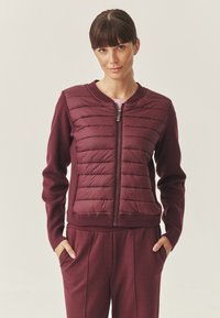 Giacca bordeaux con davanti trapuntato e polsini a costine. Presenta una zip completa e una texture morbida a maglia sulle maniche e sul retro.