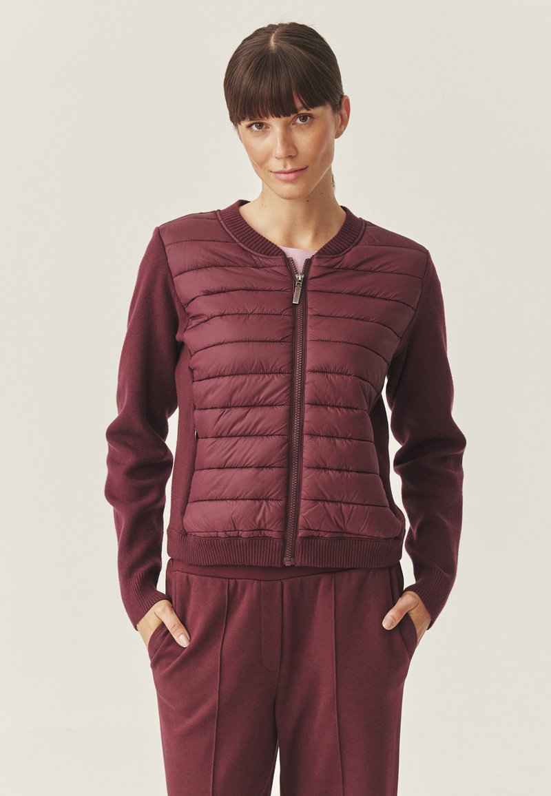 Giacca bordeaux con davanti trapuntato e polsini a costine. Presenta una zip completa e una texture morbida a maglia sulle maniche e sul retro.